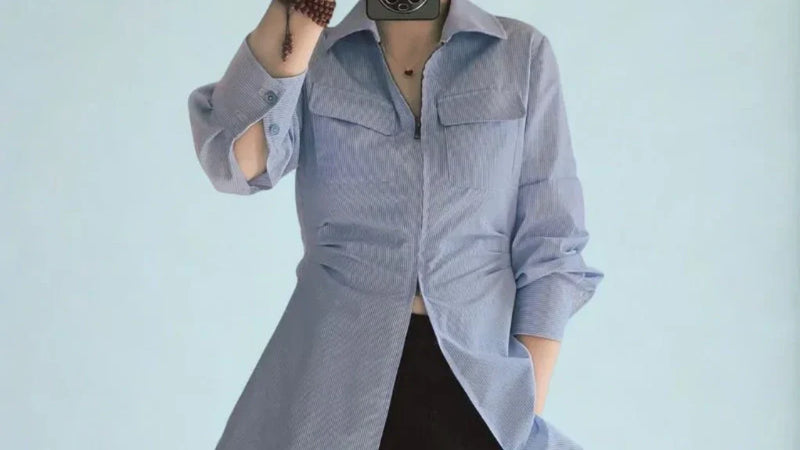 Femme portant un chemisier zippé pour femme à rayures bleu clair, manches retroussées, debout contre un fond neutre, main dans la poche et chemisier entrouvert laissant apparaître un vêtement noir en dessous.