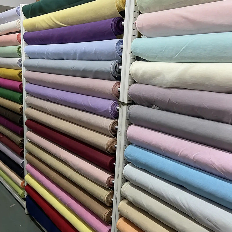 Un tissu transparent blanc cassé est drapé sur des rouleaux de tissu aux couleurs vives disposés en rangée, y compris des nuances de bleu, de vert, de turquoise, de violet, de magenta, de rose et de rouge.
