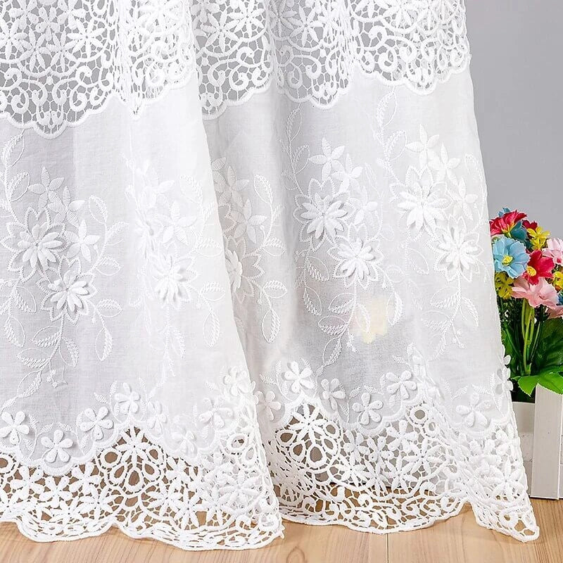 Rideau de dentelle blanche avec broderie florale complexe et bords festonnés, suspendu devant un mur gris clair. Une petite boîte de fleurs artificielles colorées est posée sur le sol à droite.