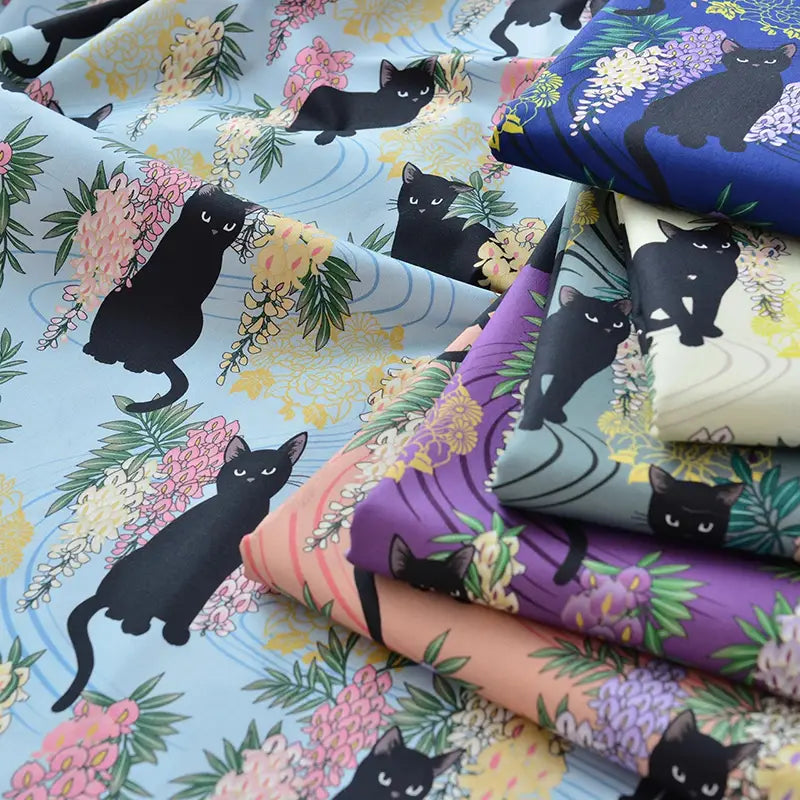 Plusieurs pièces de tissu présentant un motif répétitif de chats noirs assis parmi des fleurs roses, jaunes et blanches sur des fonds bleus, violets, crème et pêche sont empilées et étalées.
