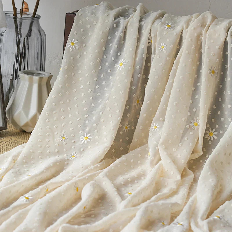 Un tissu beige clair avec un motif texturé en pointillés et de petites fleurs brodées blanches et jaunes est drapé sur une surface en bois à côté de deux vases en verre.