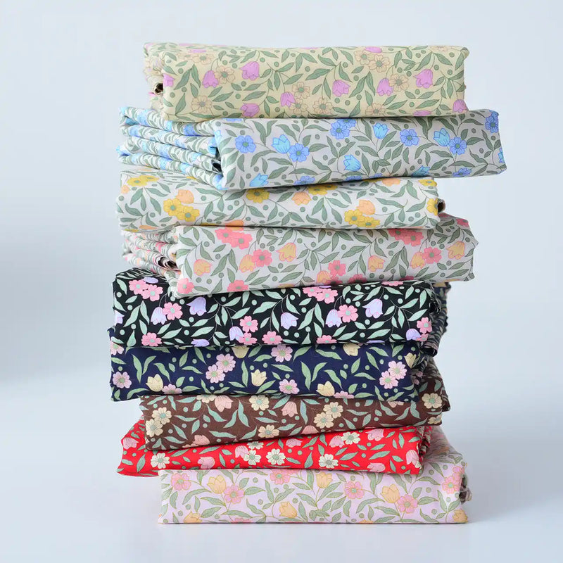 Une pile de tissus soigneusement pliés avec des motifs floraux de différentes couleurs, y compris des nuances de beige, de vert, de bleu, de rose, de noir, de marron et de rouge, est disposée sur un fond clair uni.