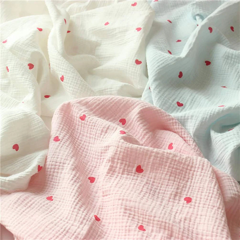 Des mousselines assorties de couleur blanche, bleu clair et rose clair, chacune ornée de petits cœurs rouges, sont disposées sur une surface plane, froissées et se chevauchant.