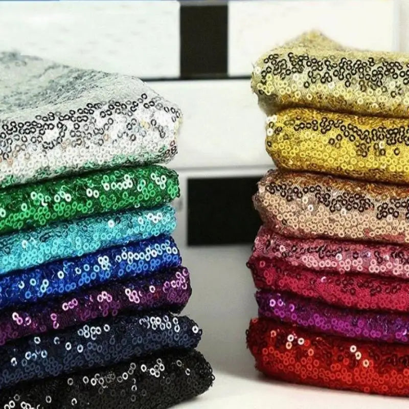 Des piles de tissus à paillettes soigneusement pliés et de différentes couleurs (argent, or, vert, bleu, violet, rose et rouge) sont disposées côte à côte sur une surface à l'arrière-plan flou.