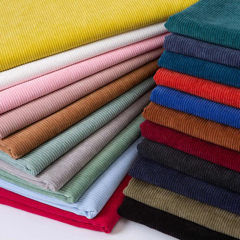 Une pile de tissus pliés en velours côtelé de différentes couleurs, notamment jaune, rose, blanc, marron, bleu clair, vert, rouge, bleu, marine, gris et noir, disposés en piles régulières et bien ordonnées.
