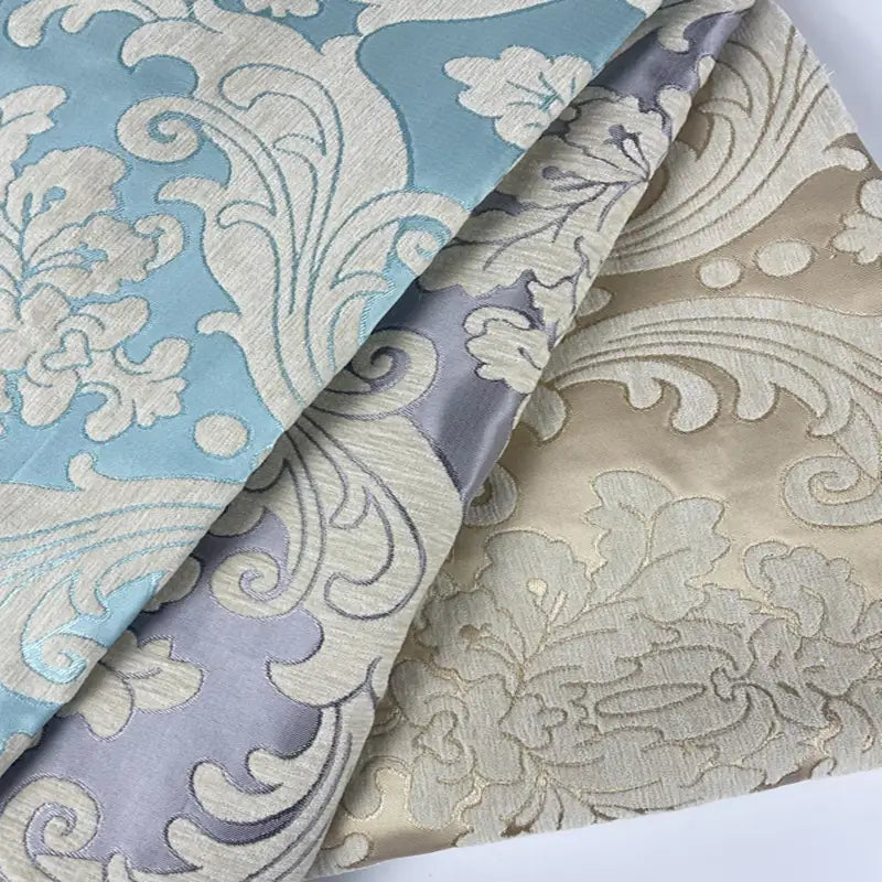 Trois pièces de tissu aux motifs floraux et feuillus ornés et en relief, dans les tons bleu, gris argenté et beige, disposées sur une surface plane et se chevauchant les unes les autres.