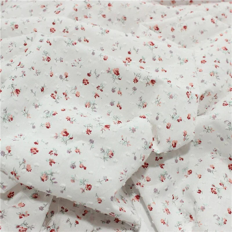 Tissu blanc avec de petits imprimés floraux rouges, roses et verts, présentant de légers plis et de la texture. Les fleurs sont dispersées uniformément sur le tissu, créant ainsi un aspect délicat et à motifs.
