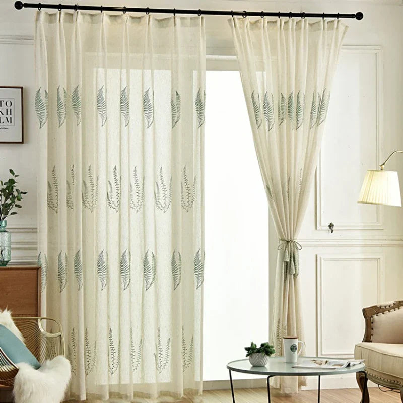 Des rideaux transparents beiges avec un motif de feuilles sont suspendus à une tringle à rideaux noire devant une grande fenêtre. Un panneau est attaché sur le côté. La pièce comprend une lampe, des chaises et une petite table avec une tasse.