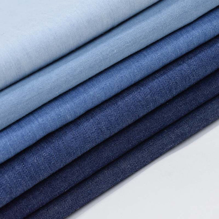 Une pile de Tissu jean 100 % coton au mètre, denim léger 4,5 oz bleu clair de Longan Craft France, plié en différentes nuances de bleu sur une surface blanche - idéal pour votre prochain projet de couture.