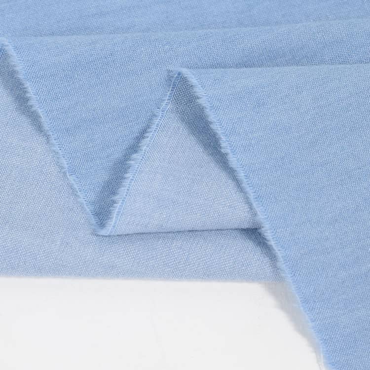 Un gros plan de Tissu jean 100 % coton au mètre denim léger 4,5 oz bleu clair de Longan Craft France montre des bords bruts et effilochés et un pli en zigzag sur une surface blanche.