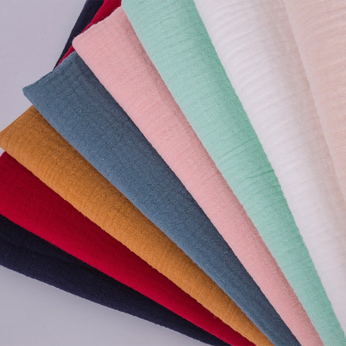 Des pièces rectangulaires du tissu double gaze 100 % coton au mètre de Longan Craft France sont disposées en éventail, montrant un tissu texturé et respirant en bleu foncé, rouge, jaune moutarde, bleu-gris, rose, vert, crème et blanc sur un fond clair.
