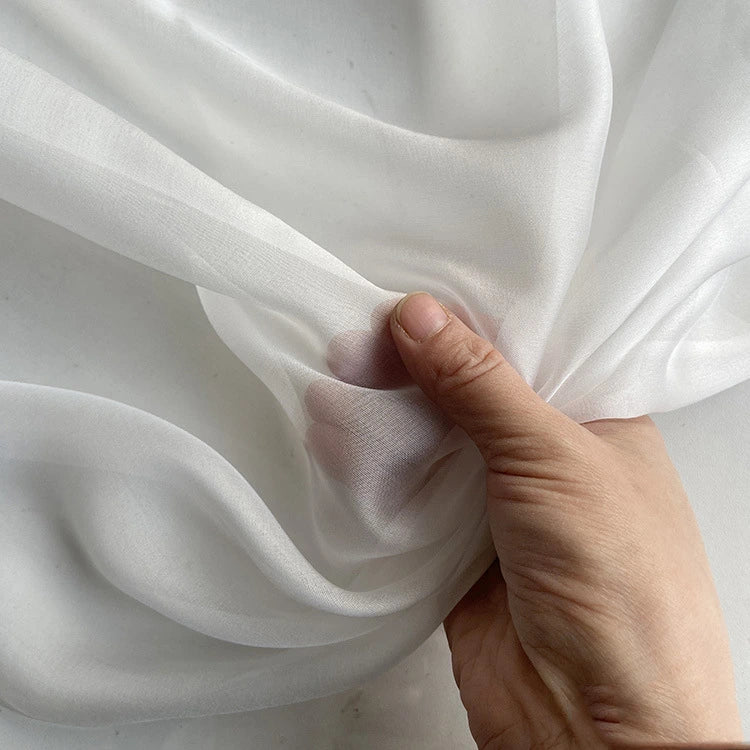 Une main fronce une Mousseline de soie de mûrier de luxe au mètre de Longan Craft France, mettant en valeur sa texture translucide et délicate - un tissu naturel et léger idéal pour la confection de vêtements.