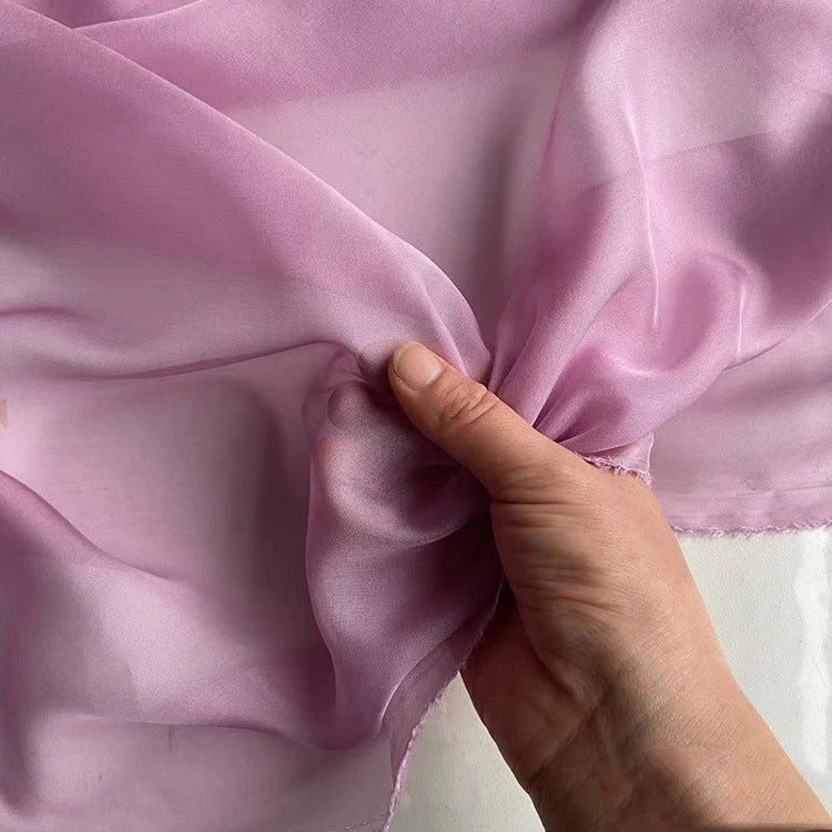 Une main pince délicatement la mousseline de soie de mûrier de luxe au mètre de Longan Craft France, un tissu naturel, léger et pur pour l'habillement, révélant sa texture lisse, sa qualité translucide et sa brillance subtile.