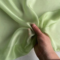 Une main tient un morceau de Mousseline de soie de mûrier de luxe au mètre, vert clair et semi-transparent, de Longan Craft France. Ce tissu naturel, léger et de première qualité semble lisse et légèrement brillant, parfait pour les vêtements.