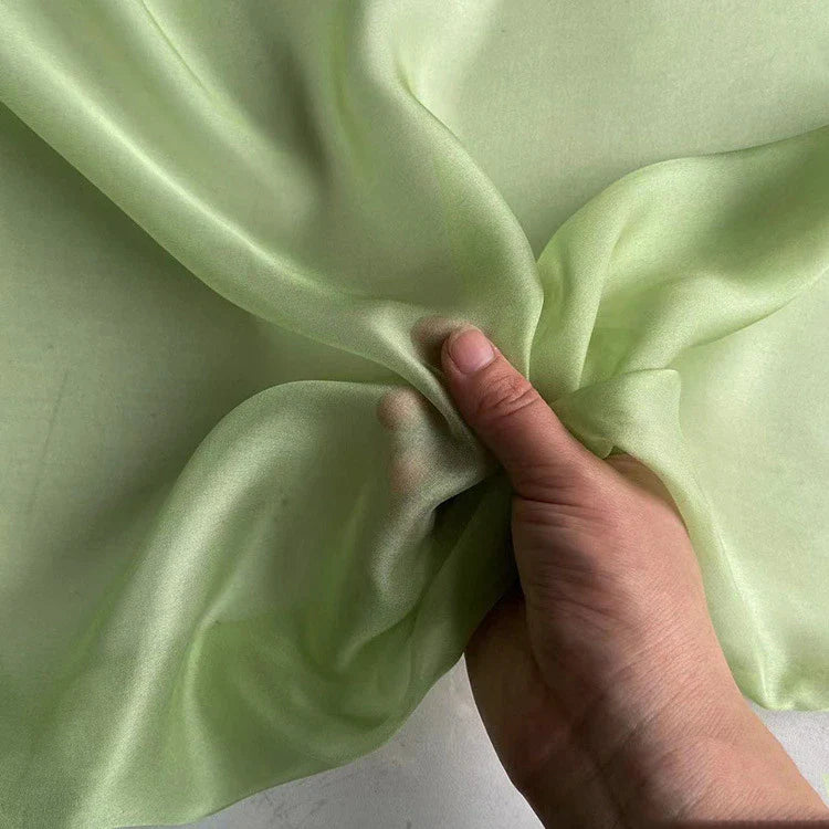 Une main tient un morceau de Mousseline de soie de mûrier de luxe au mètre, vert clair et semi-transparent, de Longan Craft France. Ce tissu naturel, léger et de première qualité semble lisse et légèrement brillant, parfait pour les vêtements.