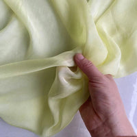 Une main tient un morceau de Mousseline de soie de mûrier de luxe au mètre de Longan Craft France, jaune pâle, sur un fond clair, mettant en valeur ce tissu naturel et léger, parfait pour l'habillement.