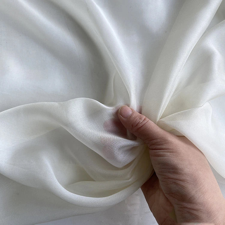 Une main tient la Mousseline de soie de mûrier de luxe au mètre de Longan Craft France sur un fond neutre, mettant en valeur la texture naturelle, légère et pure de ce tissu de robe de luxe.