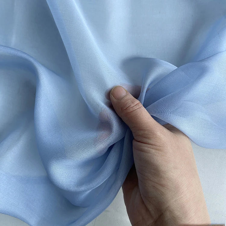 Une main tient et rassemble la Mousseline de soie de mûrier de luxe de Longan Craft France, une mousseline de soie bleue naturelle et légère destinée à l'habillement, sur fond blanc.