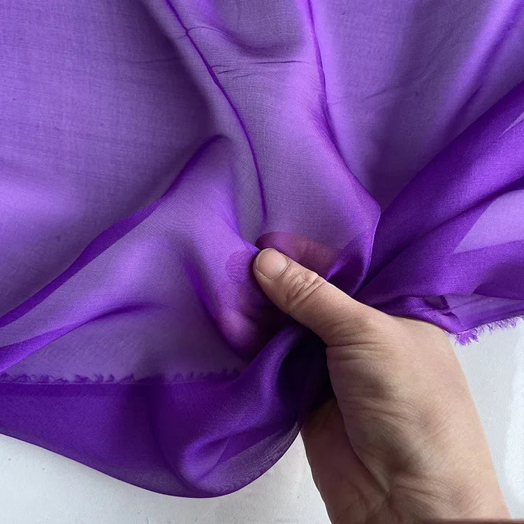 Une main tient la Mousseline de soie de mûrier violette de Longan Craft France, un tissu de soie naturelle pure, luxueux et léger, parfait pour l'habillement. La matière apparaît légèrement transparente sur un fond uni.