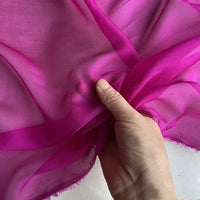 Une main tient et froisse la luxueuse mousseline de soie de mûrier magenta de Longan Craft France : un tissu léger, transparent et naturel aux bords effilochés, parfait pour la confection de vêtements.