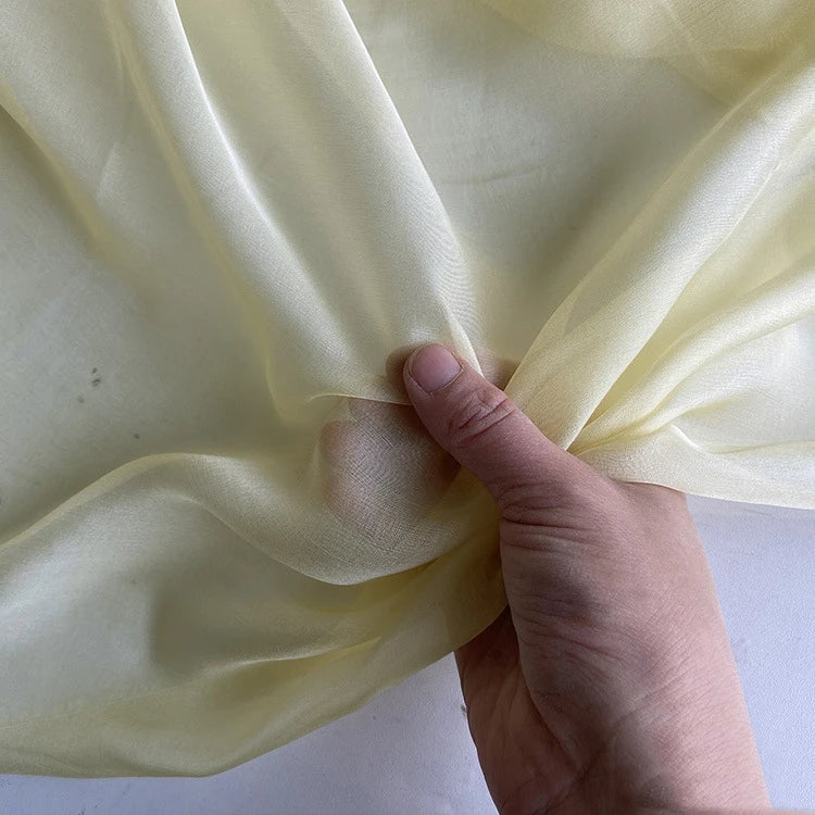 Une main rassemble un morceau de Mousseline de soie de mûrier de luxe au mètre, jaune clair et semi-transparent, de Longan Craft France - une mousseline de soie naturelle, légère et pure, idéale pour les vêtements de luxe.