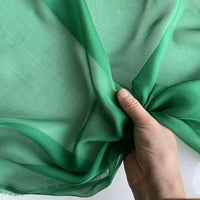 Une main rassemble un morceau de Mousseline de soie de mûrier de luxe au mètre de Longan Craft France, une mousseline de soie de mûrier naturelle vert clair, montrant sa texture délicate et translucide sur un fond flou.