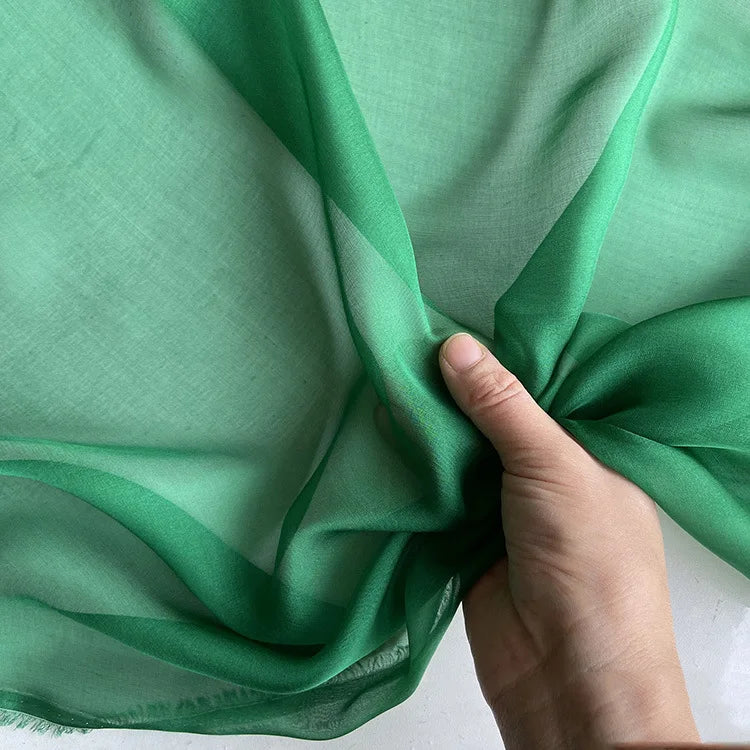 Une main rassemble un morceau de Mousseline de soie de mûrier de luxe au mètre de Longan Craft France, une mousseline de soie de mûrier naturelle vert clair, montrant sa texture délicate et translucide sur un fond flou.