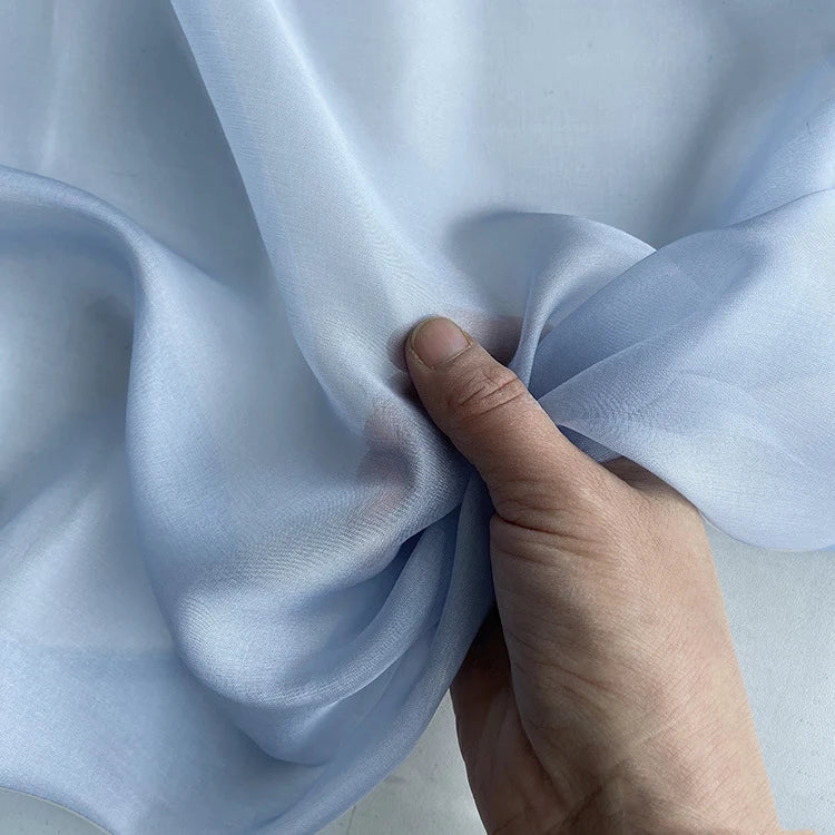 Une main rassemble un morceau de "Mousseline de soie de mûrier de luxe au mètre" bleu clair de Longan Craft France, mettant en valeur la texture délicate et légère de ce tissu en mousseline de soie naturelle, parfait pour les vêtements élégants.