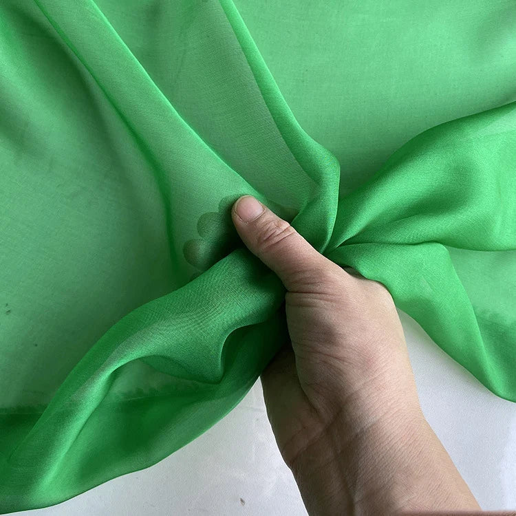 Une main tient et rassemble un morceau de Mousseline de soie de mûrier de luxe au mètre de Longan Craft France, un tissu semi-transparent et vert vif en mousseline de soie de mûrier naturel, sur un fond uni.