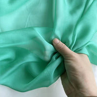 Une main fronce la Mousseline de soie de mûrier de luxe vert clair de Longan Craft France sur un fond blanc, mettant en valeur sa texture lisse et translucide - une mousseline naturelle parfaite pour les vêtements de luxe.