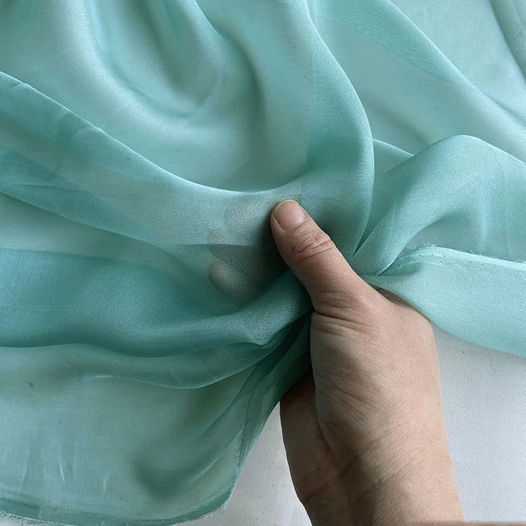 Une main tient délicatement une section de Longan Craft France Mousseline de soie de mûrier de luxe au mètre, une mousseline de soie naturelle et légère idéale pour l'habillement. La soie bleu pâle est douce, délicate et lisse, avec des plis subtils.