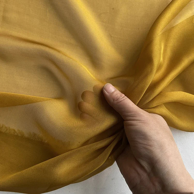 Une main tient la Mousseline de soie de mûrier de luxe au mètre de Longan Craft France - une mousseline de soie naturelle, légère et transparente, parfaite pour les vêtements de luxe. Le tissu en pure soie de mûrier apparaît semi-transparent sur un fond uni.