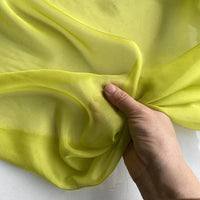 Une main tient un morceau de Longan Craft France Mousseline de soie de mûrier de luxe au mètre, une mousseline de soie jaune-vert naturelle, légère et transparente, idéale pour les vêtements élégants, sur un fond uni.