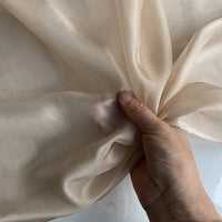 Une main fronce la Mousseline de soie de mûrier de luxe au mètre, beige clair de Longan Craft France - une mousseline de soie naturelle, légère et pure - sur un fond assorti. La translucidité de l'étoffe met en valeur sa délicatesse.
