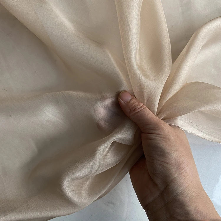 Une main fronce la Mousseline de soie de mûrier de luxe au mètre, beige clair de Longan Craft France - une mousseline de soie naturelle, légère et pure - sur un fond assorti. La translucidité de l'étoffe met en valeur sa délicatesse.