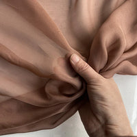 Une main saisit délicatement la luxueuse mousseline de soie de mûrier de Longan Craft France, un tissu brun clair, transparent, doux et lisse, idéal pour les vêtements élégants, vendu au mètre en tant que matériau naturel et léger.