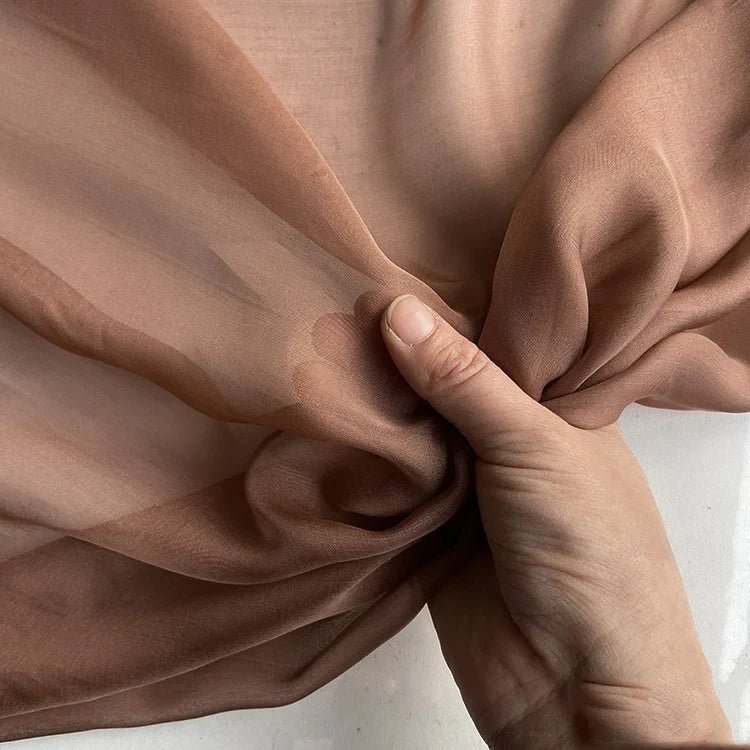 Une main saisit délicatement la luxueuse mousseline de soie de mûrier de Longan Craft France, un tissu brun clair, transparent, doux et lisse, idéal pour les vêtements élégants, vendu au mètre en tant que matériau naturel et léger.