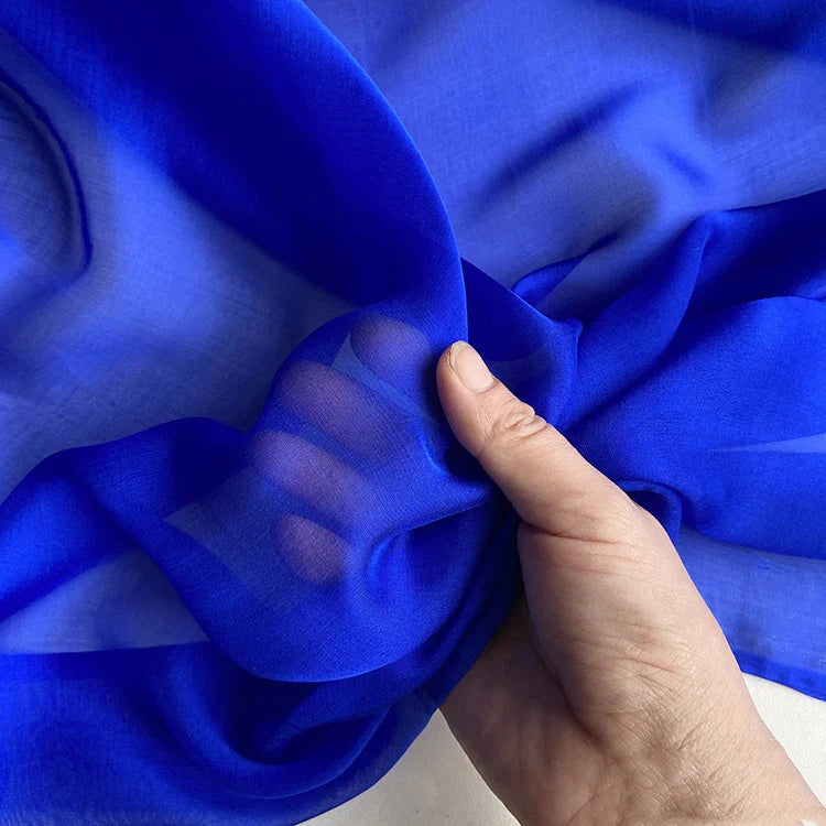 Une main rassemble un morceau de la luxueuse mousseline de soie de mûrier de Longan Craft France, un tissu naturel, léger et pur d'un bleu éclatant - parfait pour les vêtements élégants - sur un fond neutre.