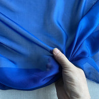 La Mousseline de soie de mûrier de luxe au mètre de Longan Craft France, bleu vif, est froncée à la main sur une surface texturée blanc cassé ; le tissu en mousseline de soie naturelle est léger et légèrement transparent.
