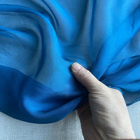 Une main tient un morceau de "Mousseline de soie de mûrier de luxe au mètre" bleu vif de Longan Craft France, un tissu transparent et léger en mousseline de soie de mûrier naturel, sur un fond blanc texturé.