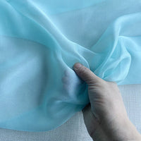 Une main tient une Mousseline de soie de mûrier de luxe au mètre bleu clair de Longan Craft France sur un fond neutre ; la mousseline de soie naturelle est transparente et légère, révélant la main grâce à sa texture délicate.