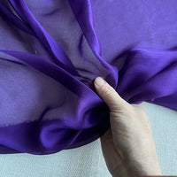 Une main tient une section de Mousseline de soie de mûrier de luxe au mètre de Longan Craft France, un tissu de soie naturelle et légère pour l'habillement, présenté sur un fond légèrement texturé.
