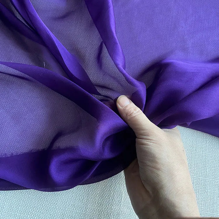 Une main tient une section de Mousseline de soie de mûrier de luxe au mètre de Longan Craft France, un tissu de soie naturelle et légère pour l'habillement, présenté sur un fond légèrement texturé.
