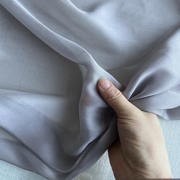 Une main soulève la luxueuse mousseline de soie de mûrier de Longan Craft France, montrant sa texture lisse, légère et légèrement transparente - idéale pour les vêtements élégants - sur un fond neutre.