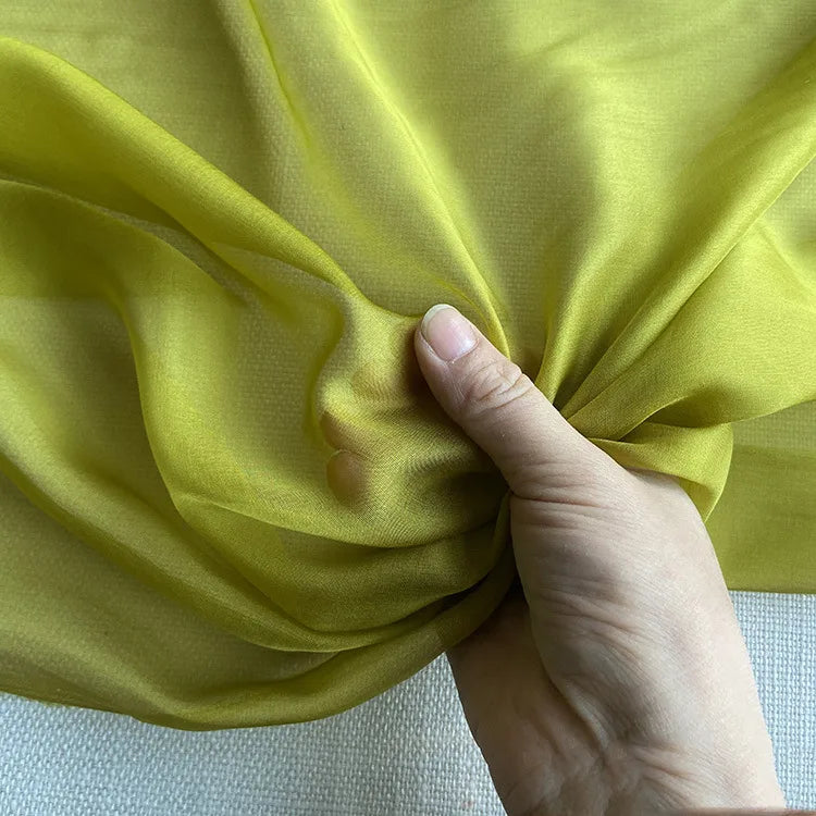 Une main rassemble un morceau de la luxueuse mousseline de soie de mûrier de Longan Craft France, une mousseline naturelle transparente, jaune-vert clair, vendue au mètre - délicate, translucide et parfaite pour les vêtements élégants.