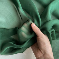 Une main tient un morceau de Mousseline de soie de mûrier de luxe au mètre de Longan Craft France, un tissu vert transparent, naturel et léger, à la texture légèrement translucide et lisse, parfait pour les vêtements élégants.