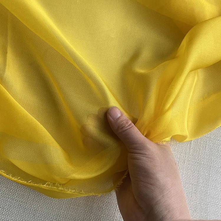 Une main présente la luxueuse mousseline de soie de mûrier au mètre de Longan Craft France, une mousseline de soie de mûrier au mètre - un tissu léger et naturel pour l'habillement, aux bords légèrement effilochés, sur un fond texturé pâle.