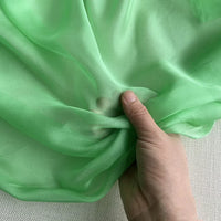Une main tient la Mousseline de soie de mûrier de luxe au mètre de Longan Craft France - une fine mousseline de soie de mûrier semi-transparente parfaite pour les vêtements de luxe, présentée sur un fond texturé neutre.