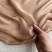 Une main tient la luxueuse mousseline de soie de mûrier de Longan Craft France, un tissu vestimentaire naturel, léger et pur au mètre, montrant sa transparence, sa douceur et ses bords subtilement effilochés sur une surface légère.