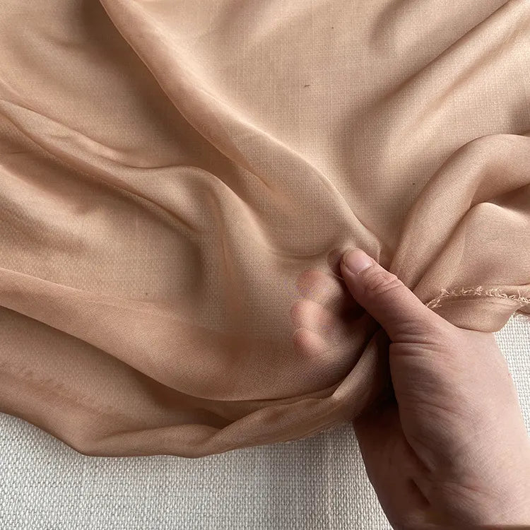 Une main tient la luxueuse mousseline de soie de mûrier de Longan Craft France, un tissu vestimentaire naturel, léger et pur au mètre, montrant sa transparence, sa douceur et ses bords subtilement effilochés sur une surface légère.